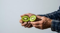 Senior male farmer with weathered hands holding freshly cut kiwi fruit halves, displaying green flesh and seeds against neutral background for agricultural and food production concepts.