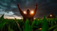 Man with raised arms standing in green cornfield at dramatic sunset, silhouette against moody sky with glowing sun and dark storm clouds creating spiritual atmosphere.