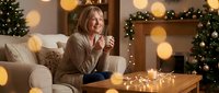 Smiling blonde woman in cozy sweater enjoying hot drink on sofa in festively decorated living room with Christmas tree and warm bokeh lights.