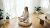 Young woman of East Asian descent in casual beige loungewear sitting cross-legged on floor cushion in bright minimalist living room with potted plants and wooden furniture, practicing meditation or mindfulness.