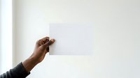 Male hand with dark skin tone holding blank white card against neutral wall background for mockup and branding purposes.