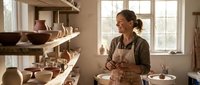 Female potter working with clay in ceramic studio workshop surrounded by handmade pottery bowls and vessels on wooden shelves with natural window light.