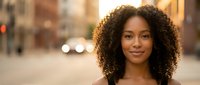 Young African American woman with curly hair smiling confidently on urban city street during golden hour sunset for lifestyle and portrait photography.