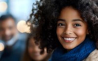 Young African American woman with natural curly hair wearing blue scarf smiling warmly at camera. Soft bokeh lights create cozy atmosphere in background.