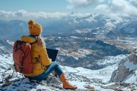 Remote worker in yellow winter jacket using laptop on snowy mountain peak, enjoying spectacular winter landscape view. Digital nomad lifestyle and outdoor adventure concept.