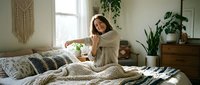 Young woman with medium-length dark hair and light skin tone, wearing a beige cardigan, sitting on a bed with neutral bedding in a bright, plant-filled bedroom. Morning light streams through a window, creating a calm, peaceful atmosphere. Ideal for lifestyle, wellness, and home decor content.