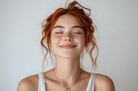 Young redhead woman with closed eyes and gentle smile expressing joy and serenity against light background, wearing white tank top and delicate necklace.
