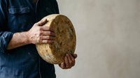 Caucasian male baker or artisan holding freshly baked round sourdough bread loaf with rustic crust, wearing denim shirt, isolated against neutral background for bakery, food business, and culinary content.