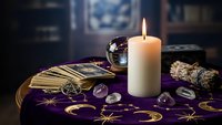 Mystical fortune telling setup with tarot cards, crystal ball, burning candle and gemstones on purple celestial cloth for spiritual guidance sessions.