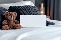 Brown teddy bear sitting on white bed sheets next to blank card mockup with dark knitted blanket, cozy bedroom interior setting for greeting or invitation design.