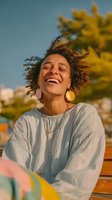 Young woman with curly reddish-brown hair and medium skin tone smiling outdoors on sunny day wearing light striped shirt and gold hoop earrings, seated on wooden bench with blurred yellow flowers and clear blue sky background.