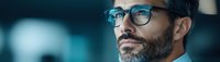 Professional middle aged man with gray beard wearing modern blue glasses looking thoughtfully into distance against dark background with soft lighting.