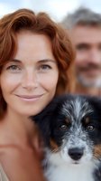 Smiling redhead woman with freckles and green eyes posing with mixed breed dog while mature man stands in background. Close up portrait showing joy and companionship.