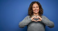 Smiling middle-aged Caucasian woman making heart shape with hands wearing gray sweater on blue background for wellness and self-care campaigns.