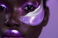 Close up of African American woman with creative purple makeup, metallic lipstick, and luminous white face paint detail in artistic beauty portrait with dark lighting.