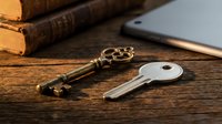 Antique brass key with ornate handle and modern silver key on wooden desk surface next to vintage books and tablet for security, access, and mystery concepts.