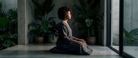 Young woman with curly brown hair, appearing to be of mixed ethnicity, sits in meditation pose on concrete floor in modern minimalist space with potted plants and large windows, practicing mindfulness and wellness.