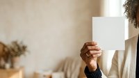 Unrecognizable person holding blank white card in front of face indoors, wearing neutral tones for mockup and branding purposes.