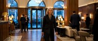 Professional blonde businesswoman in elegant black suit standing confidently in upscale hotel lobby with arched windows and sophisticated interior design.
