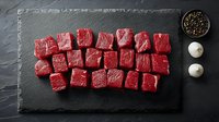 Fresh raw beef cubes arranged on dark slate board with peppercorns and garlic for cooking preparation and culinary presentations.