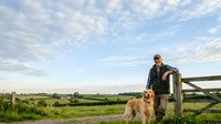 Man in casual attire standing with golden retriever by wooden fence in rural countryside landscape with rolling fields and blue sky.