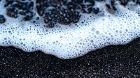 White foam and bubbles on black volcanic sand beach creates dramatic contrast in abstract macro photography composition. Natural texture background.