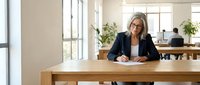 Middle-aged woman with light skin tone and blonde hair wearing glasses and dark blazer writing at wooden desk in bright office with plants and colleagues working in background.