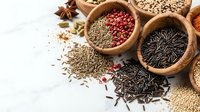 Assorted spices and seeds in wooden bowls including black rice, lentils, fennel, and peppercorns arranged on white background for culinary cooking ingredients.