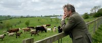 Middle-aged Caucasian farmer in green jacket talking on phone while leaning on wooden fence overlooking pasture with grazing cattle herd in countryside.