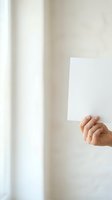 Hand holding blank white card against neutral wall with natural light from window, suitable for mockups, advertising, and promotional campaigns.