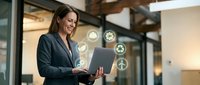 Businesswoman holding laptop with digital icons floating around her in modern office environment, representing technology, connectivity, and professional growth.