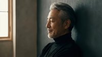 Mature Asian man with gray hair in black turtleneck leaning against wall in contemplative pose with natural window light creating peaceful indoor atmosphere.
