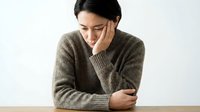 Young Asian woman in cozy brown sweater sitting at wooden table with hand on cheek, expressing contemplation and introspection in minimalist setting.