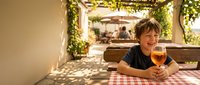 Young boy enjoying orange juice at outdoor restaurant terrace with vine-covered pergola and checkered tablecloth on sunny summer day for family dining concepts.