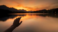 Hand reaching toward golden sunset over calm mountain lake with perfect reflections creating peaceful meditation moment.