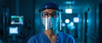 Healthcare worker in protective face shield and mask standing in hospital corridor with blue lighting for medical safety and pandemic protection concepts.