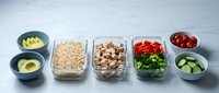 Healthy meal prep containers with fresh vegetables, nuts, grains and fruits arranged on white surface for weekly planning and nutritious eating habits.
