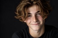 Young Caucasian male teenager with wavy blonde hair smiling warmly at camera against dark background. Natural portrait lighting creates authentic mood.