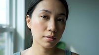 Young Asian woman with natural makeup looking thoughtfully at camera in soft natural light by window for wellness and lifestyle content.
