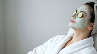 Young woman relaxing with green facial mask and cucumber slices on eyes wearing white bathrobe during spa treatment for skincare and wellness routine.