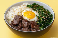 Korean beef bowl with marinated meat, sunny side up egg, rice noodles and sauteed spinach garnished with sesame seeds on yellow background.