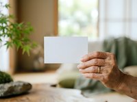 Senior adult hand holding blank white card in zen garden setting with potted plants and natural window light.