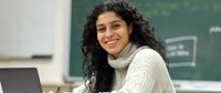 Young female teacher with curly dark hair and cream sweater smiling while working on laptop in classroom with chalkboard background. Professional education setting.