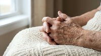 Elderly hands clasped together in prayer or meditation, showing wrinkled skin and age spots on cream knitted blanket by window, conveying peace and spirituality.