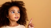 Young African American girl with natural curly hair pointing upward with curious expression against warm beige background, looking at copy space above.
