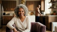 Happy mature African American woman with curly gray hair smiling warmly in modern kitchen while family cooks in background for lifestyle content.