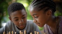 Young African American couple playing chess outdoors, smiling while concentrating on game strategy. Natural lighting and bokeh background create intimate atmosphere.