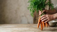 Male hand holding fresh organic carrots with green leafy tops on wooden surface against neutral beige wall background for cooking and healthy eating content.