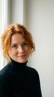 Young redhead woman with curly hair wearing black turtleneck sweater smiling at camera in bright modern interior with natural window light.