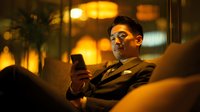 Asian businessman relaxing on couch using smartphone in warm evening lighting at modern home interior for lifestyle and technology concepts.